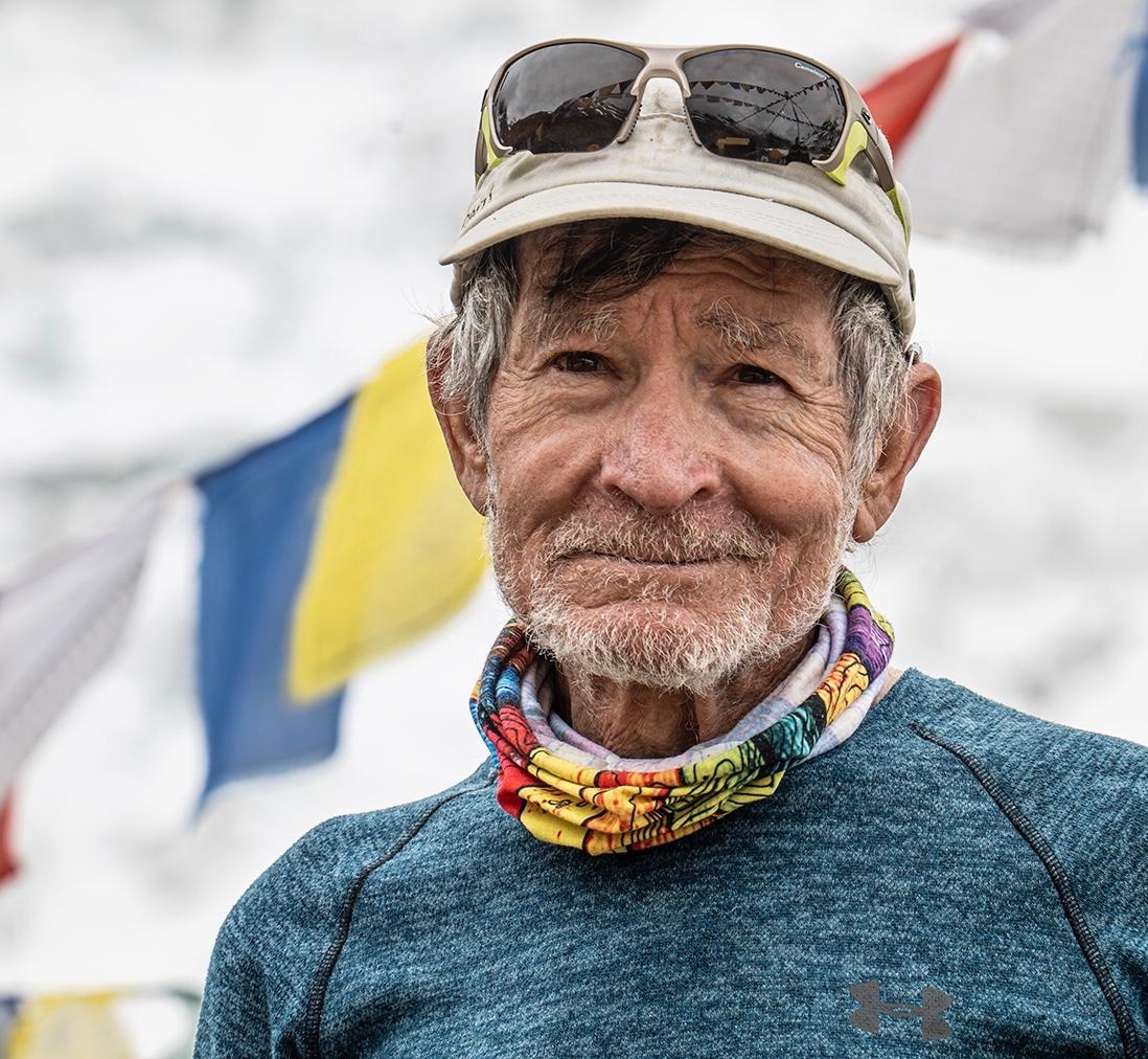 Retrato de Carlos Soria con gorra y braga de colores, frente a banderas de oración